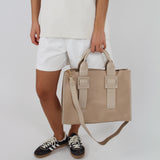 En person iført hvid T-shirt og shorts bærer en beige taske i hånden. Personen står i sort-hvide sneakers på en hvid baggrund.