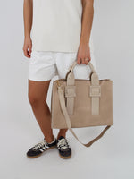 En person iført hvid T-shirt og shorts bærer en beige taske i hånden. Personen står i sort-hvide sneakers på en hvid baggrund.