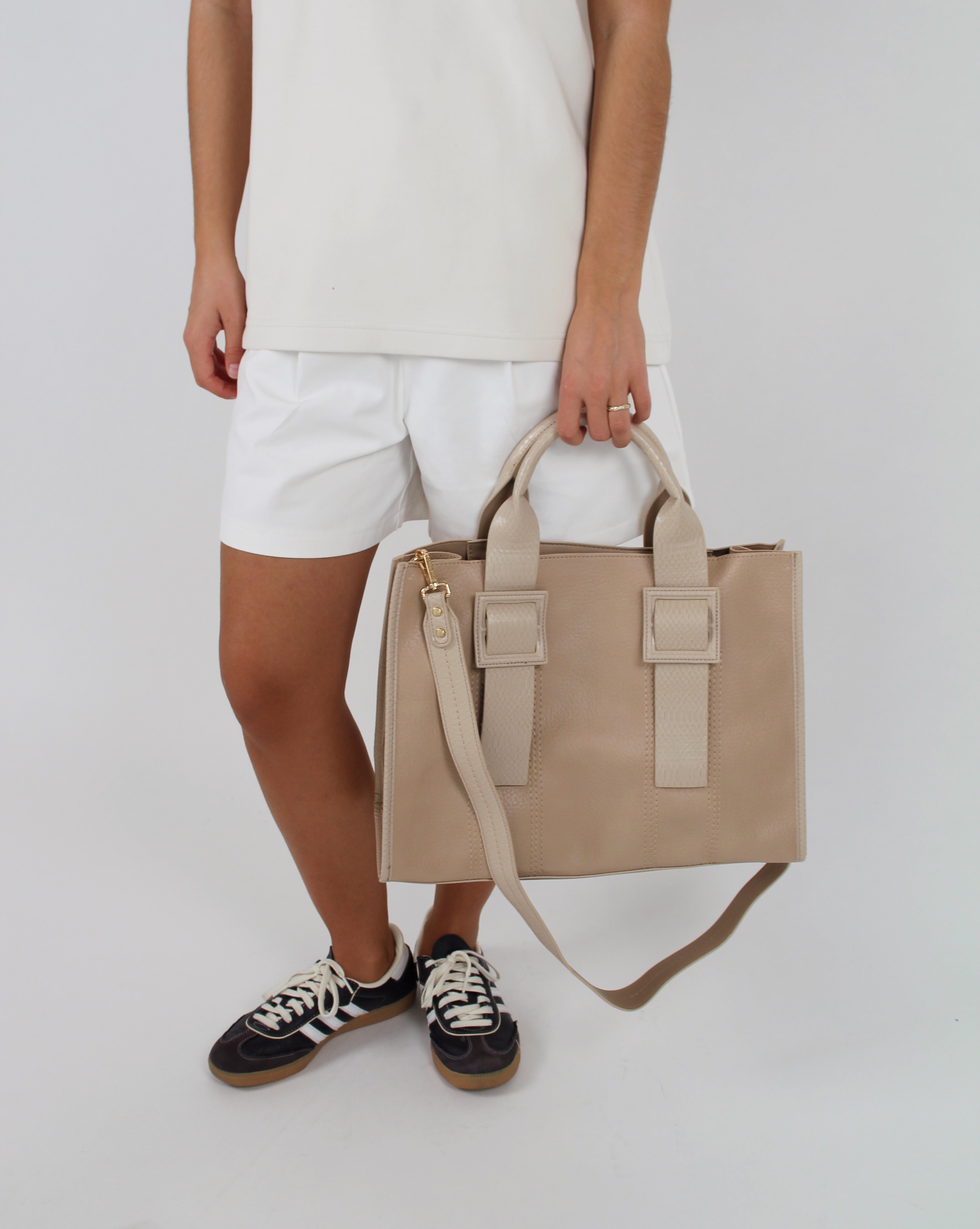 En person iført hvid T-shirt og shorts bærer en beige taske i hånden. Personen står i sort-hvide sneakers på en hvid baggrund.