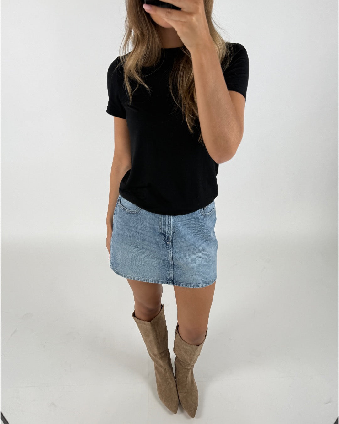 En person bærer en sort T-shirt og en blå denimnederdel, tager et spejlbillede, iført beige støvler. Baggrunden er en enkel, hvid væg.