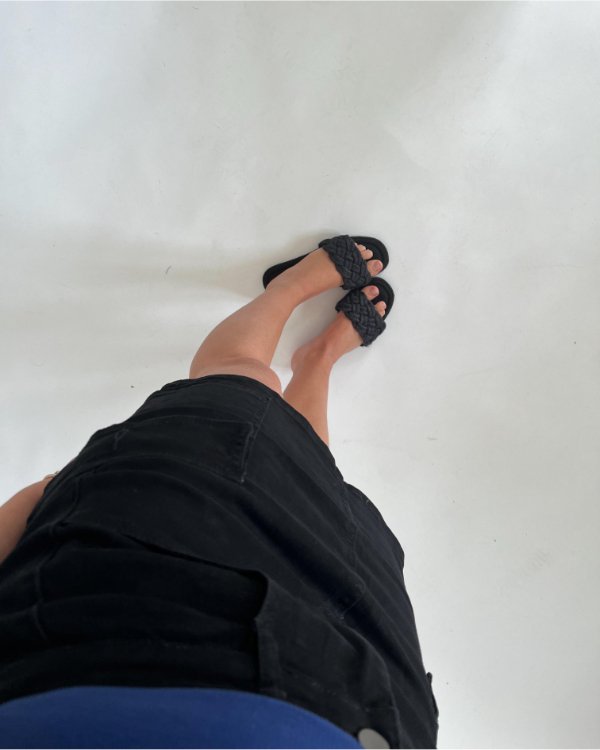 Nogle ben iført sorte sandaler står på et lyst gulv. Personen har en sort nederdel og en blå top på.