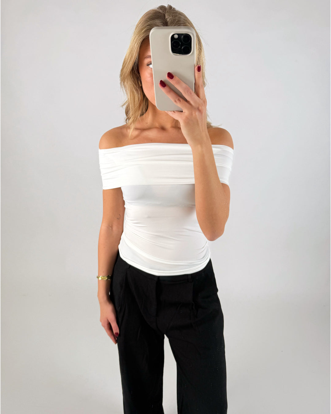 En person tager et spejlbillede iført en hvid off-shoulder top og sorte bukser. Personen holder en telefon op foran ansigtet i en minimalistisk, lys baggrund.