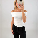 En person tager et spejlbillede iført en hvid off-shoulder top og sorte bukser. Personen holder en telefon op foran ansigtet i en minimalistisk, lys baggrund.