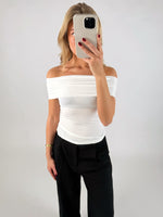 En person tager et spejlbillede iført en hvid off-shoulder top og sorte bukser. Personen holder en telefon op foran ansigtet i en minimalistisk, lys baggrund.