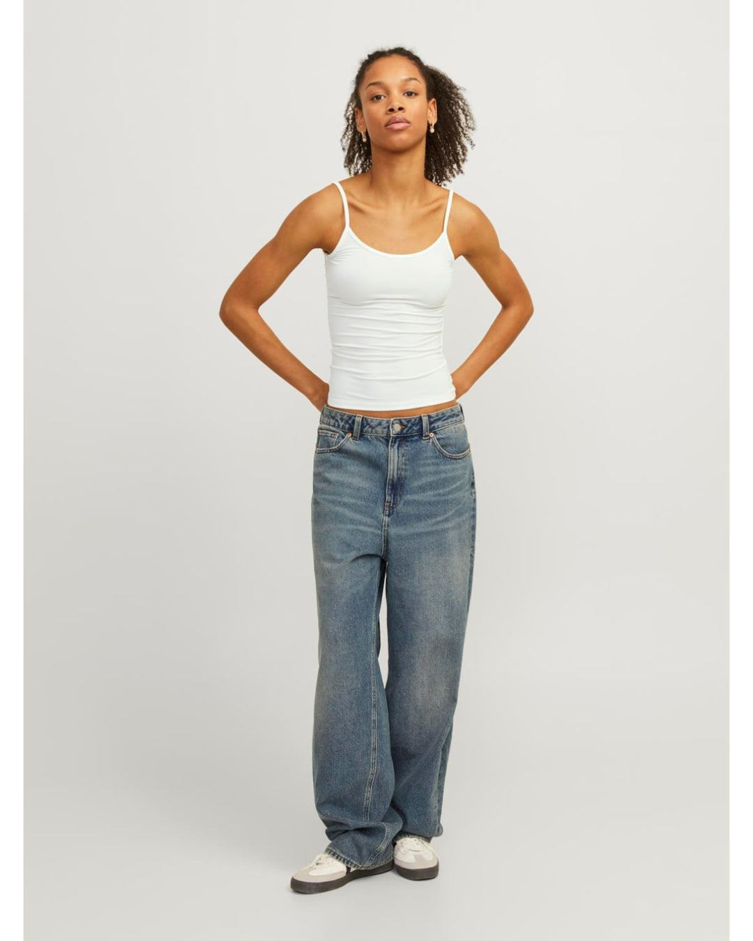 En person står med hænderne på hofterne iført en hvid tanktop og løse blå jeans. Baggrunden er grå og minimalistisk.