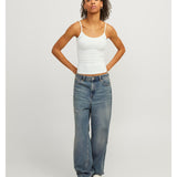 En person står med hænderne på hofterne iført en hvid tanktop og løse blå jeans. Baggrunden er grå og minimalistisk.