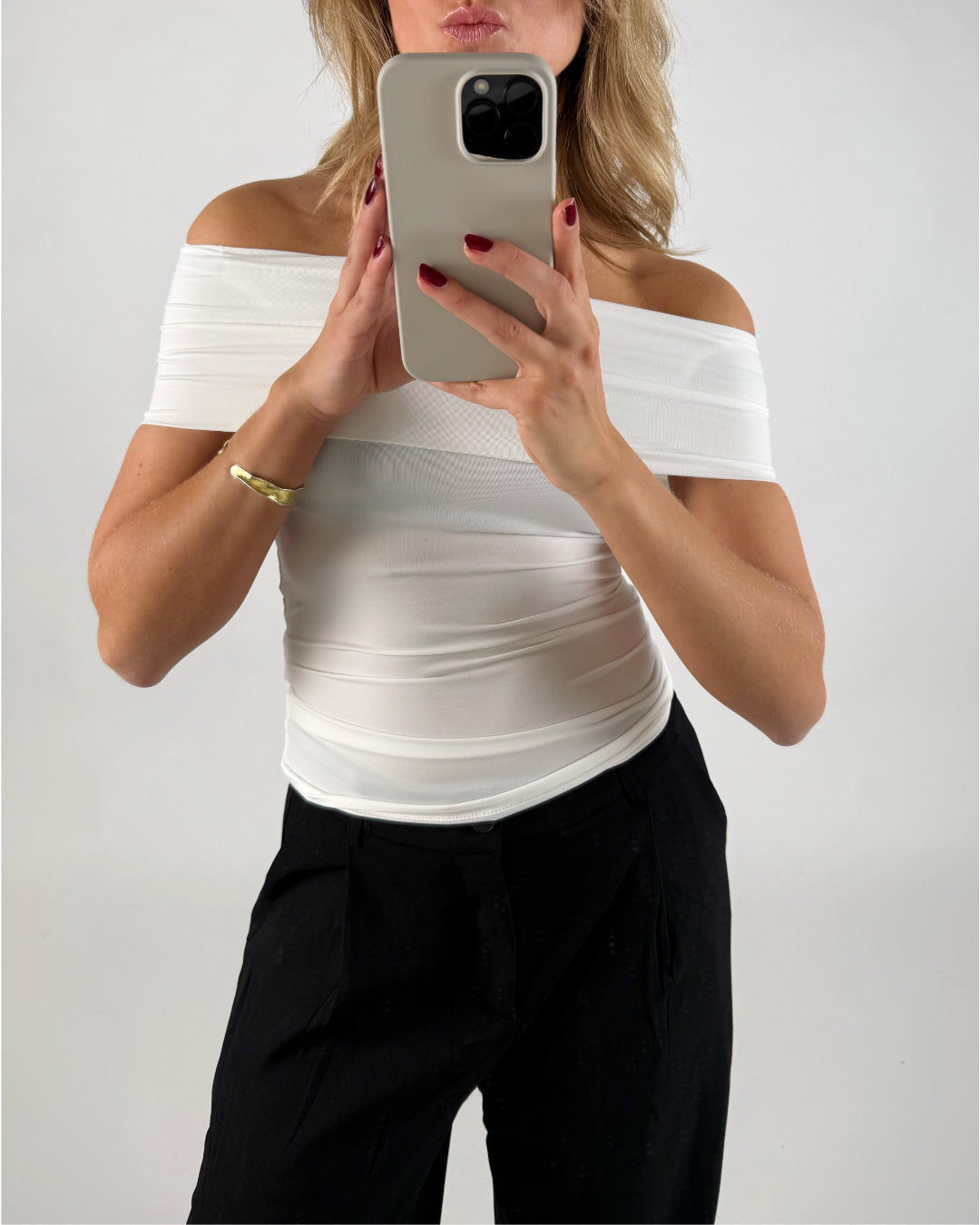 En person i en hvid top holder en mobiltelefon op foran ansigtet i en selfie-position, baggrund er neutral og lys.