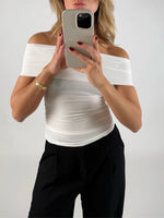 En person i en hvid top holder en mobiltelefon op foran ansigtet i en selfie-position, baggrund er neutral og lys.