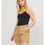 En kvinde med langt blond hår står roligt, iført sort tanktop og beige shorts. Hun hviler en hånd i lommen, med en neutral baggrund.