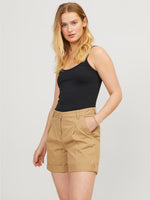 En kvinde med langt blond hår står roligt, iført sort tanktop og beige shorts. Hun hviler en hånd i lommen, med en neutral baggrund.