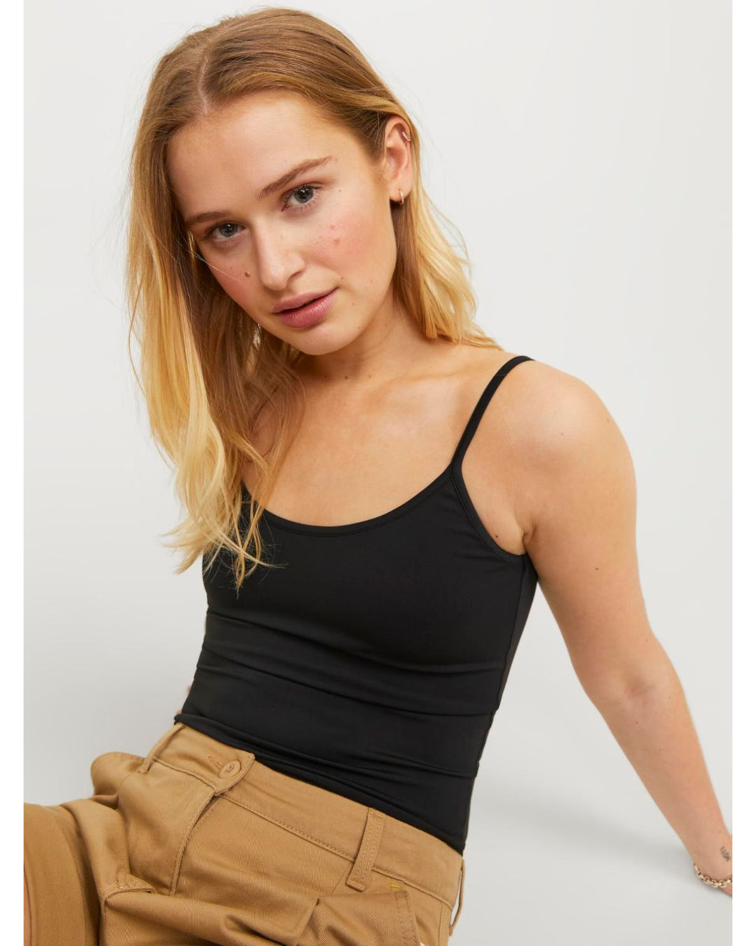 En person sidder iført en sort top og beige bukser, kigger mod kameraet. Baggrunden er enkel og lys.