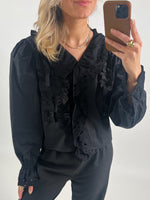En person i sort bluse med flæsedetaljer tager en selfie med en smartphone. Personen har lyst hår og bærer minimal smykker i et neutralt oplyst rum.