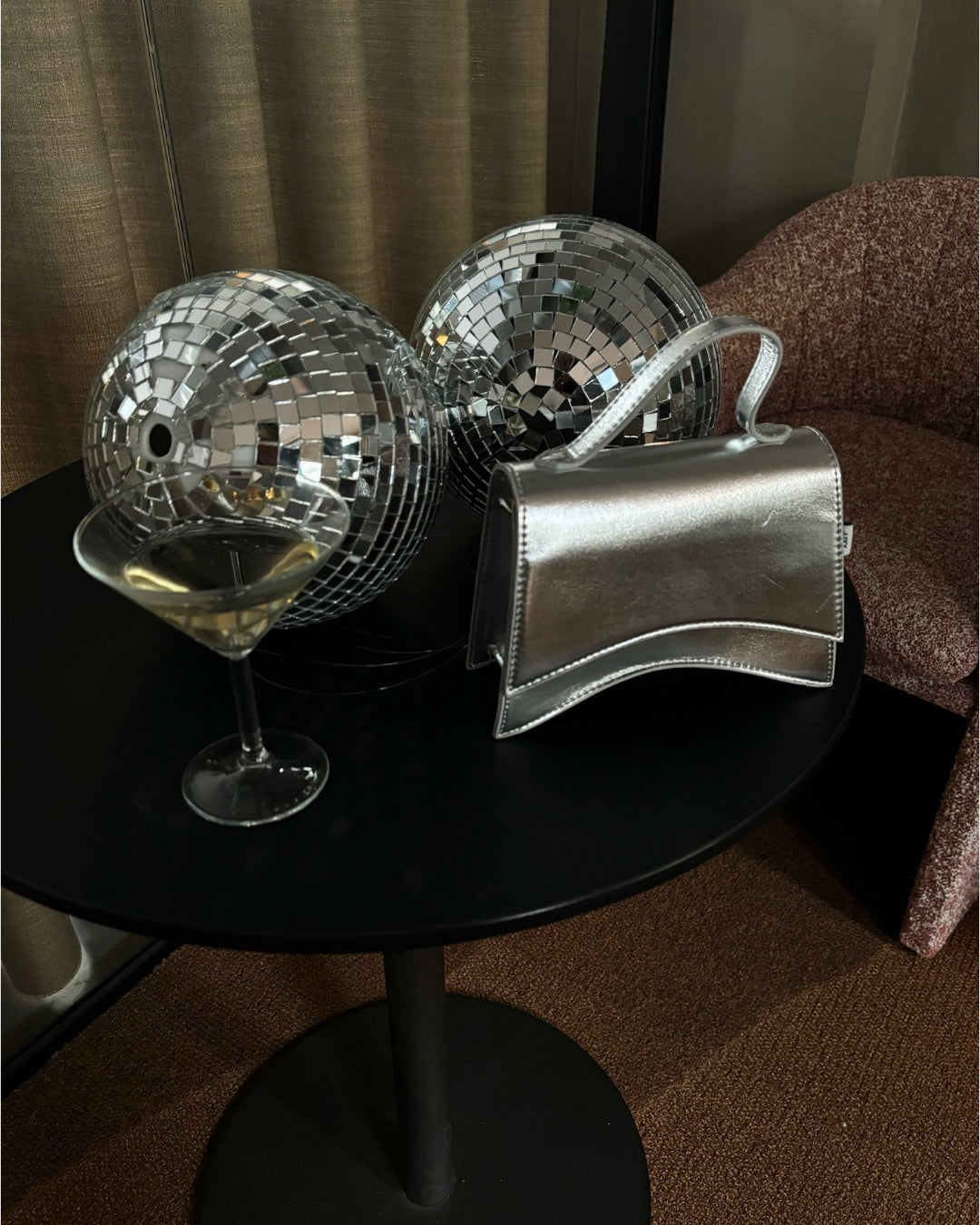 To disco-kugler og en sølvfarvet taske ligger på et rundt sort bord. Et martiniglas står ved siden af i et rum med brune stole og gardiner.