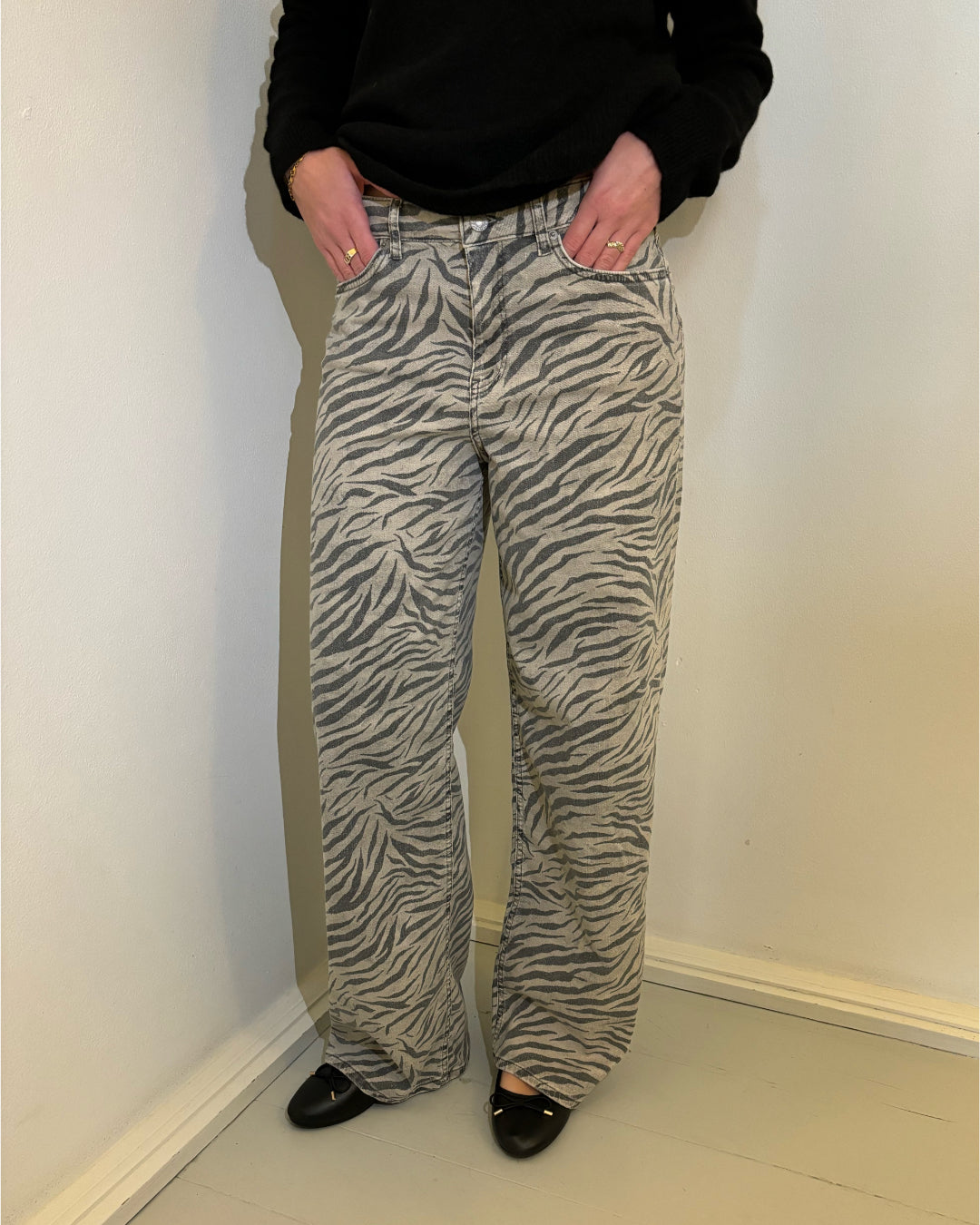 Person iført zebra-mønstrede bukser står med hænderne i lommerne i et enkelt rum med lyse vægge og gulv. Vedkommende har sorte sko og en sort trøje på.