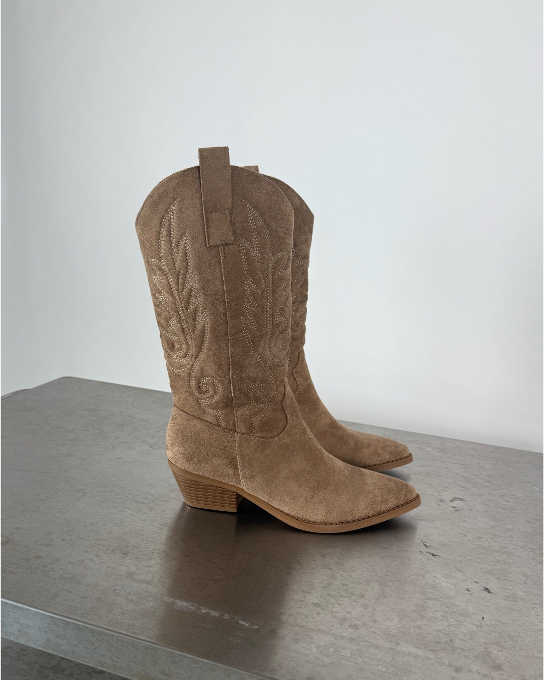 Klassiske brune cowboystøvler står stille på en grå overflade. De har broderede mønstre og er lavet af ruskind med en lav hæl. Baggrunden er en neutral, lys væg.