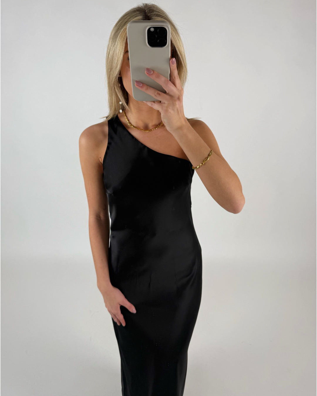 En person i en sort, ærmeløs kjole tager et spejlbillede med en smartphone. Personen bærer guldsmykker og står i et minimalistisk, hvidt rum.