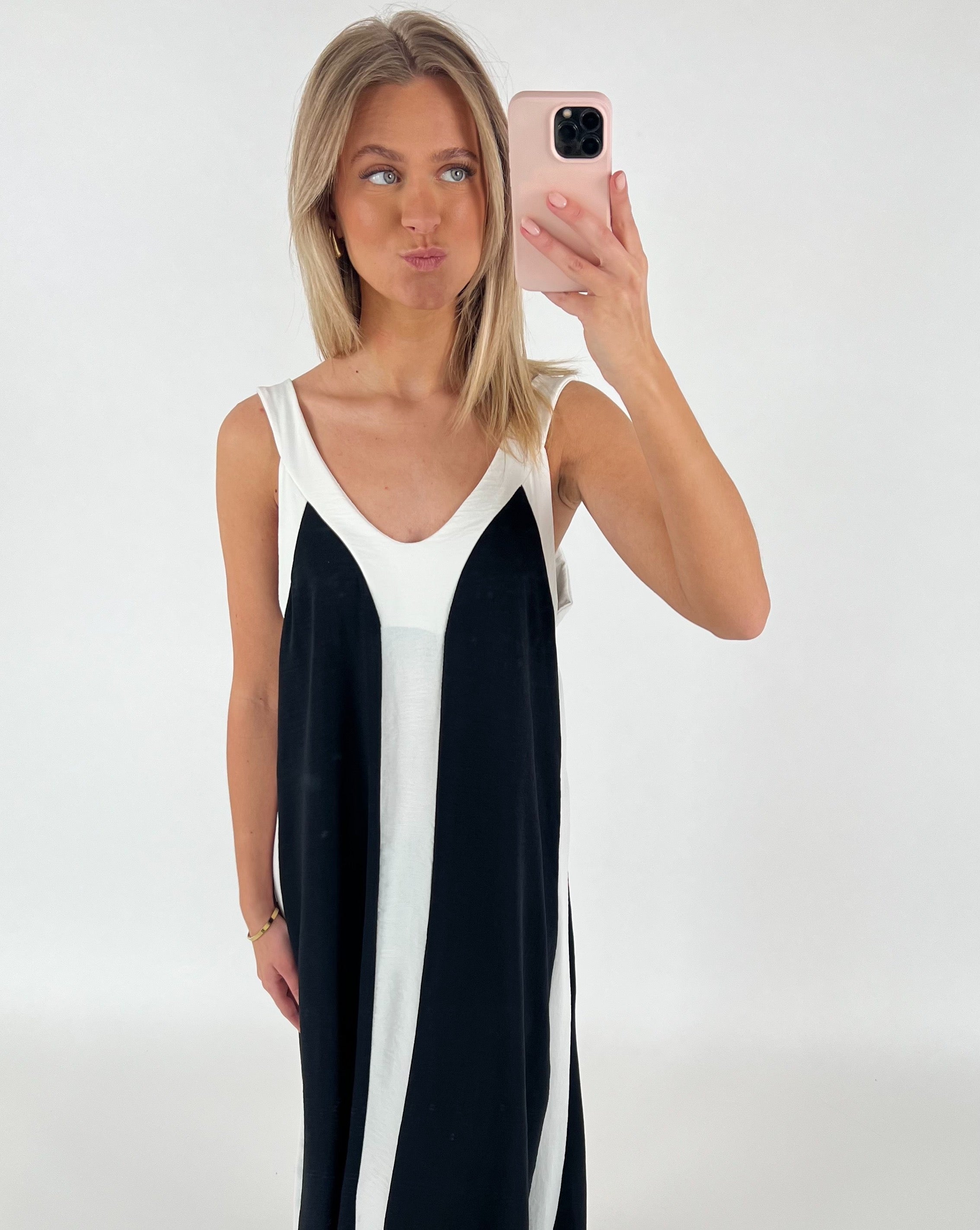 En kvinde i en sort-hvid kjole tager en selfie med en pink telefon i en lys, minimalistisk baggrund. Hun poserer med en humoristisk ansigtsudtryk.