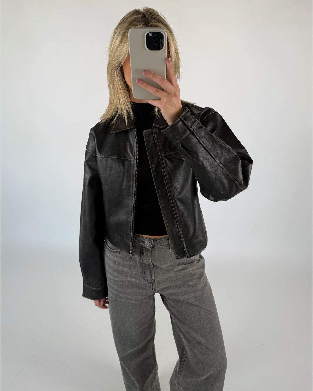 En person iført en sort læderjakke tager et spejlfoto med en smartphone i et minimalistisk, hvidt rum. Personen har lyse hår og grå jeans på.