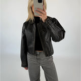 En person iført en sort læderjakke tager et spejlfoto med en smartphone i et minimalistisk, hvidt rum. Personen har lyse hår og grå jeans på.