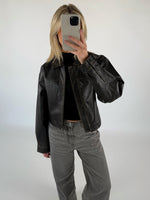En person iført en sort læderjakke tager et spejlfoto med en smartphone i et minimalistisk, hvidt rum. Personen har lyse hår og grå jeans på.