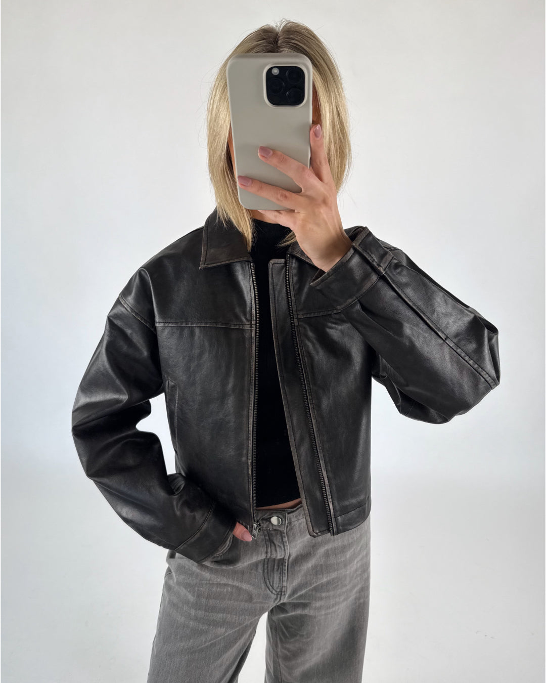 En person tager et spejlbillede, iført en sort læderjakke og grå jeans, i et lyst rum. Personen holder en mobiltelefon foran ansigtet.