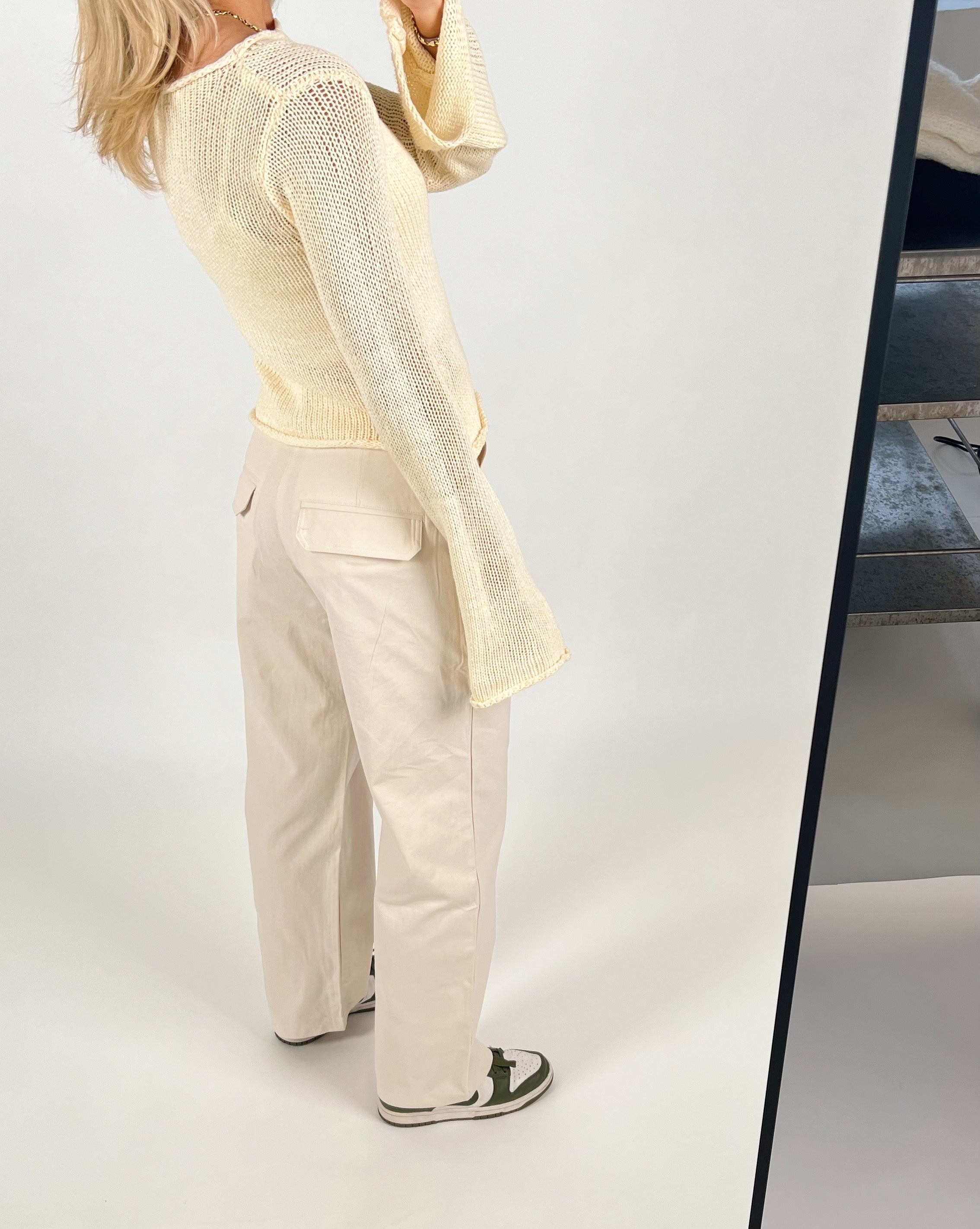 En person iført en beige trøje og cremefarvede bukser står i et lyst rum, spejlbilledet delvist synligt til højre. Personen bærer hvide sneakers med grønne detaljer.