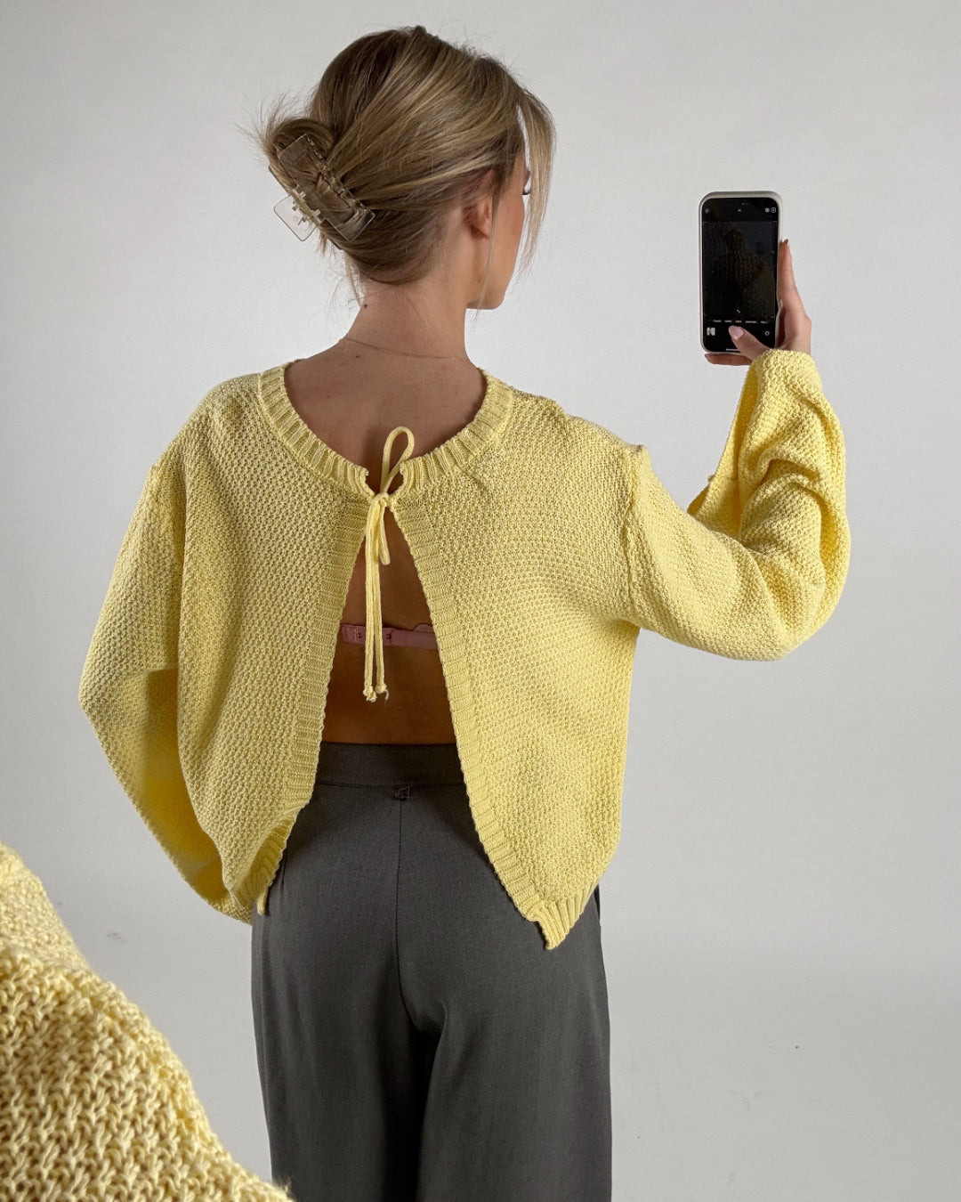 Kvinde iført gul sweater, tager et billede med telefonen. Sweateren har et åbent design med snore i nakken. Hun står foran en neutral, lys baggrund.