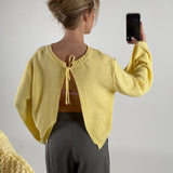 Kvinde iført gul sweater, tager et billede med telefonen. Sweateren har et åbent design med snore i nakken. Hun står foran en neutral, lys baggrund.