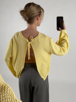 Kvinde iført gul sweater, tager et billede med telefonen. Sweateren har et åbent design med snore i nakken. Hun står foran en neutral, lys baggrund.