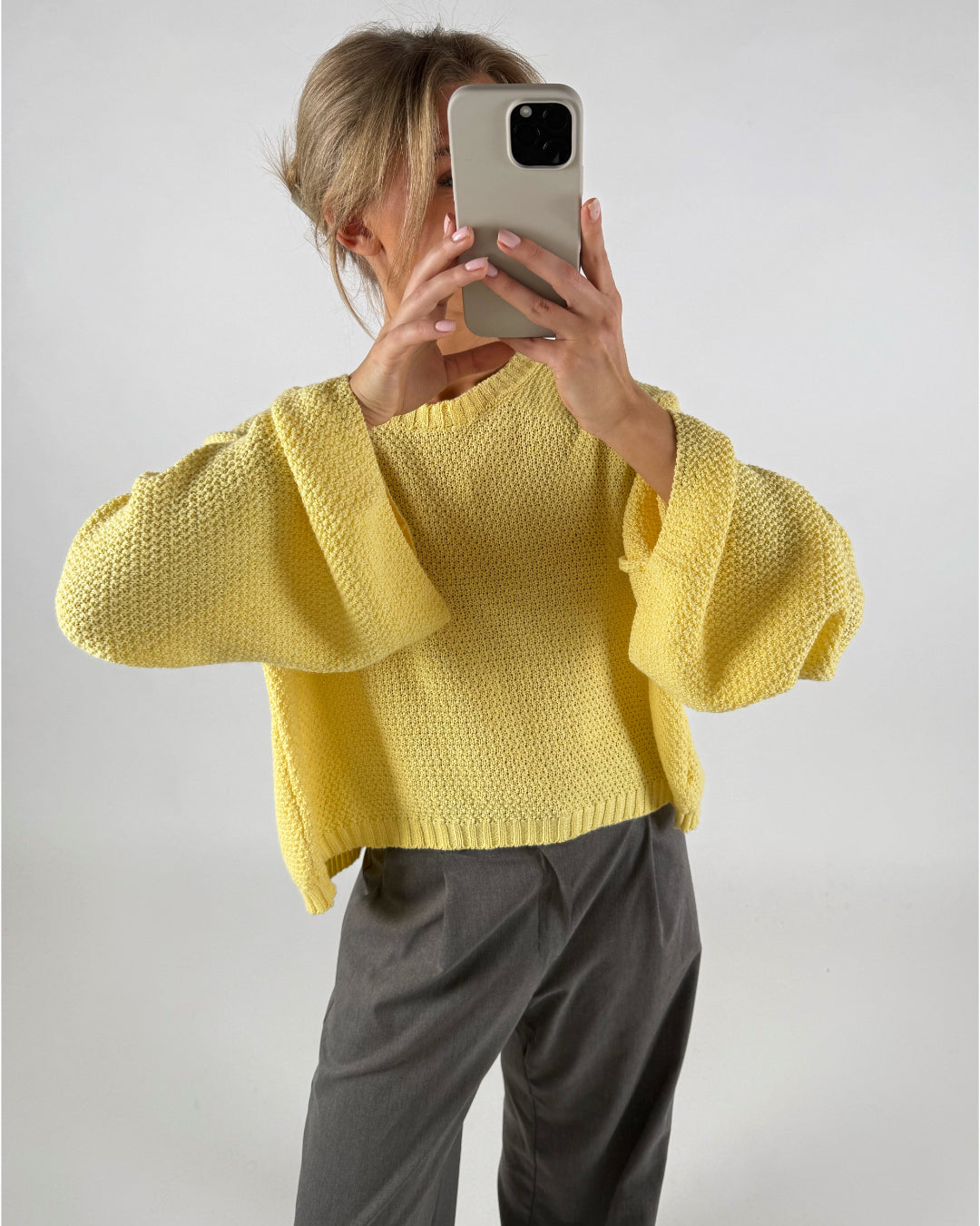 En person iført en gul sweater tager et billede med en telefon. Personen står i et neutralt rum og bærer grå bukser.