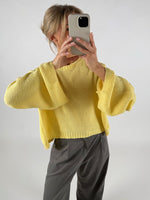 En person iført en gul sweater tager et billede med en telefon. Personen står i et neutralt rum og bærer grå bukser.