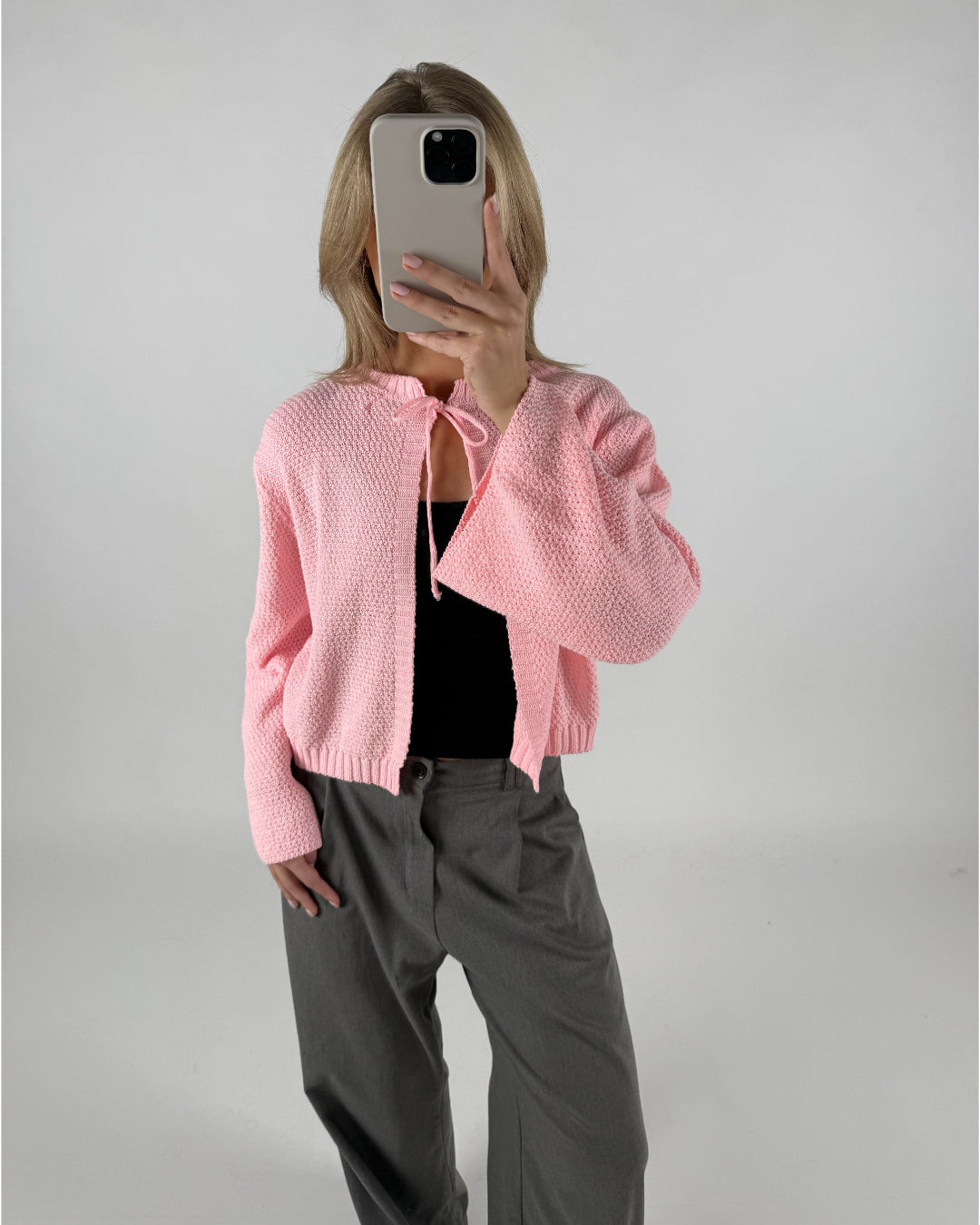 En person iført en lyserød cardigan tager et spejlbillede med en smartphone, står i et minimalistisk rum, og bærer grå bukser og sort top.