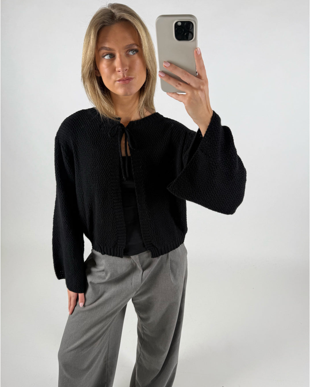 En person iført en sort jakke poser foran et spejl og tager en selfie med en smartphone, omgivet af en simpel hvid baggrund.