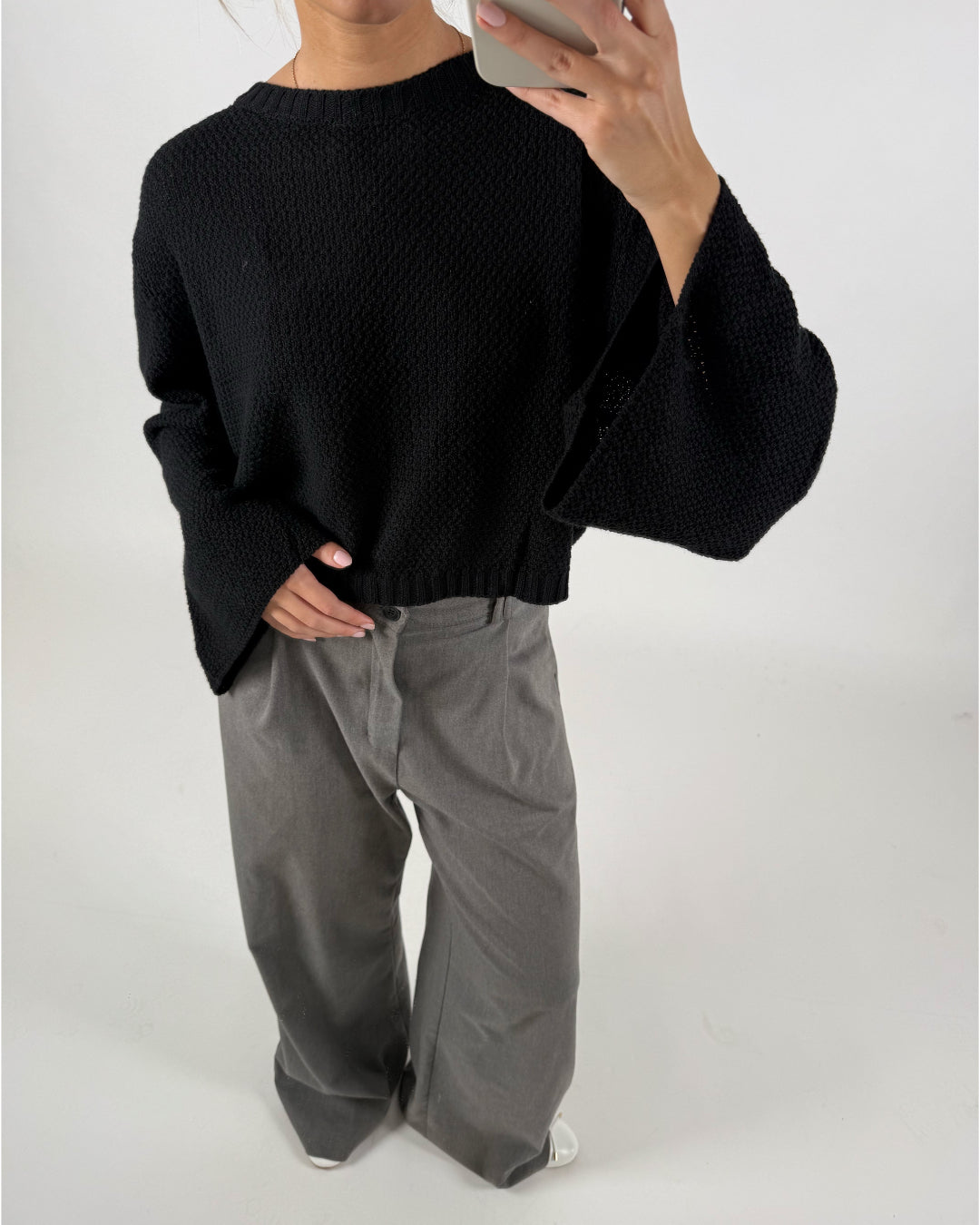 En person iført en sort, strikket sweater og grå, løse bukser holder en telefon op foran spejlet i et minimalistisk rum med hvid baggrund.