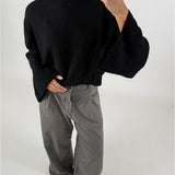En person iført en sort, strikket sweater og grå, løse bukser holder en telefon op foran spejlet i et minimalistisk rum med hvid baggrund.