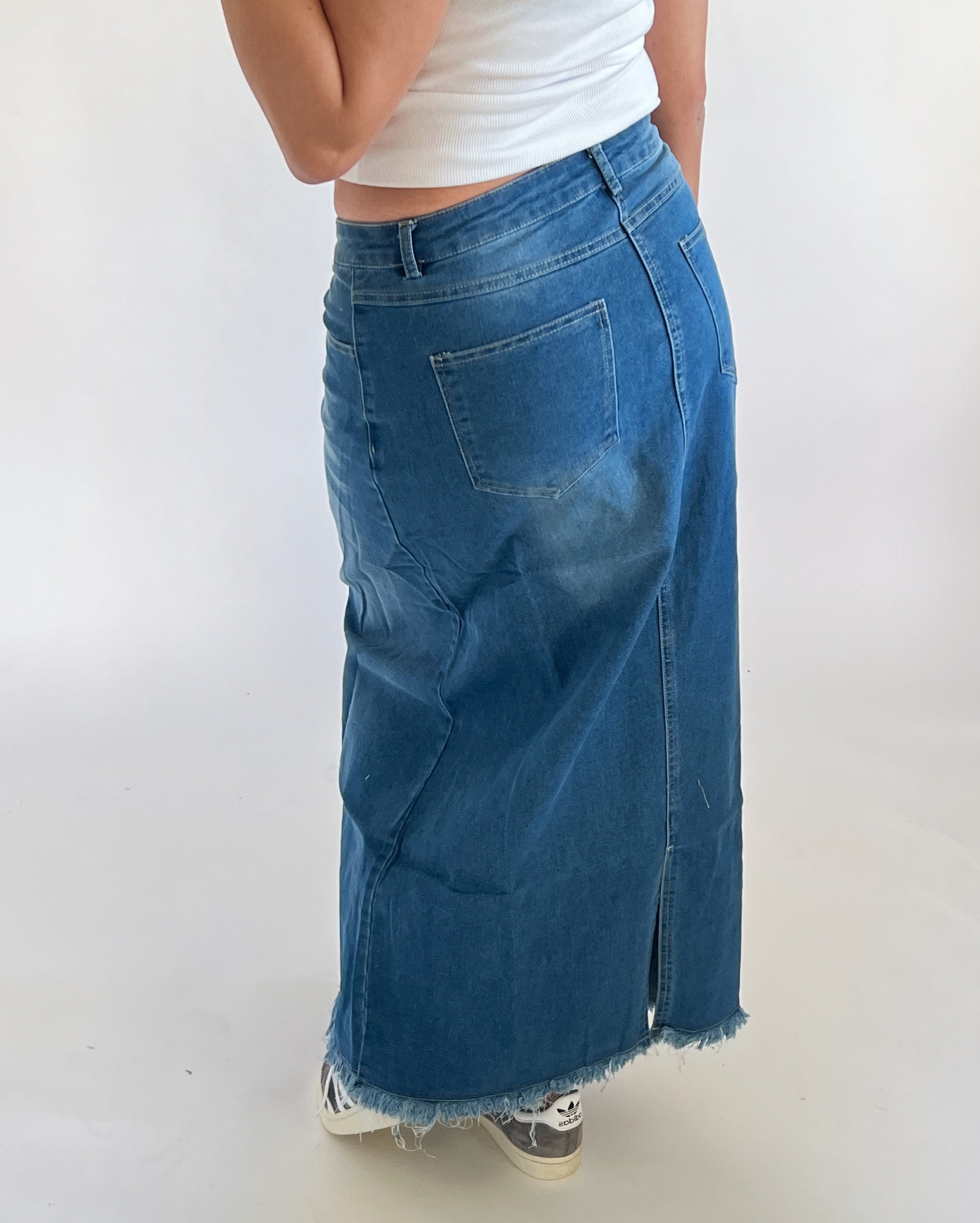 En person bærer en blue denim maxi-nederdel med frynser og en hvid top. Nederdelen har lommer og en slids bagpå. Baggrunden er en simpel, lys flade.