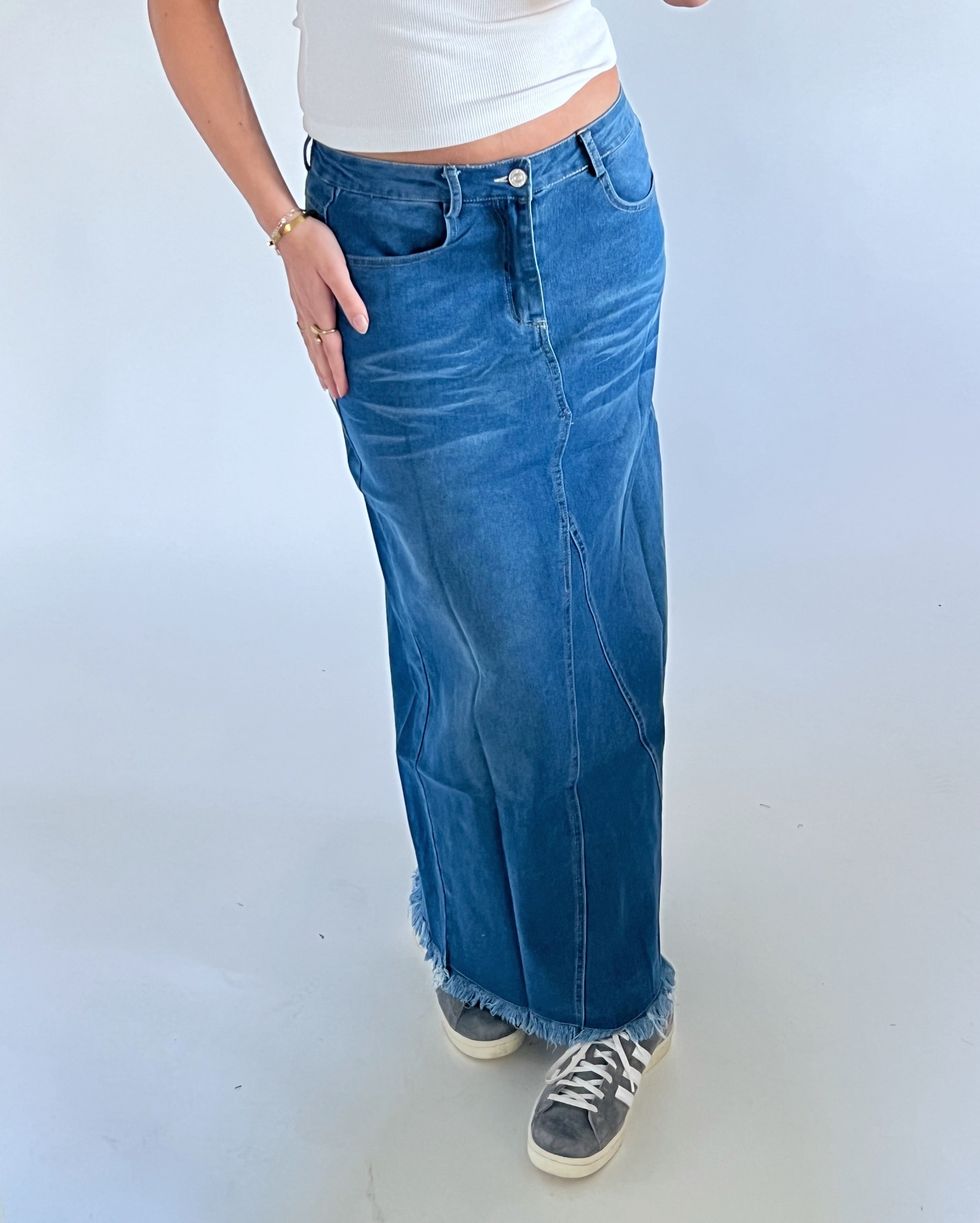 En person iført en tætsiddende, blå denimnederdel og sneakers står mod en neutral baggrund. Personen bærer en hvid top og har en hånd i lommen.
