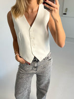 En person med lyseblondt hår bærer en hvid vest og stenvasket jeans, mens de tager en selfie i et minimalistisk rum. Personen har hånden i lommen og bærer et armbånd.