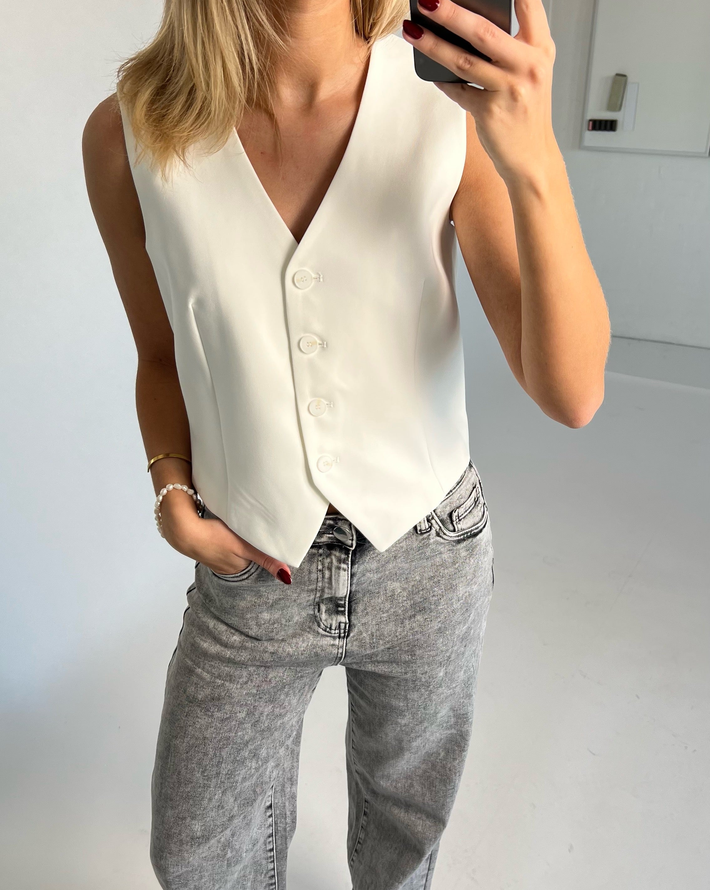 En person med lyseblondt hår bærer en hvid vest og stenvasket jeans, mens de tager en selfie i et minimalistisk rum. Personen har hånden i lommen og bærer et armbånd.