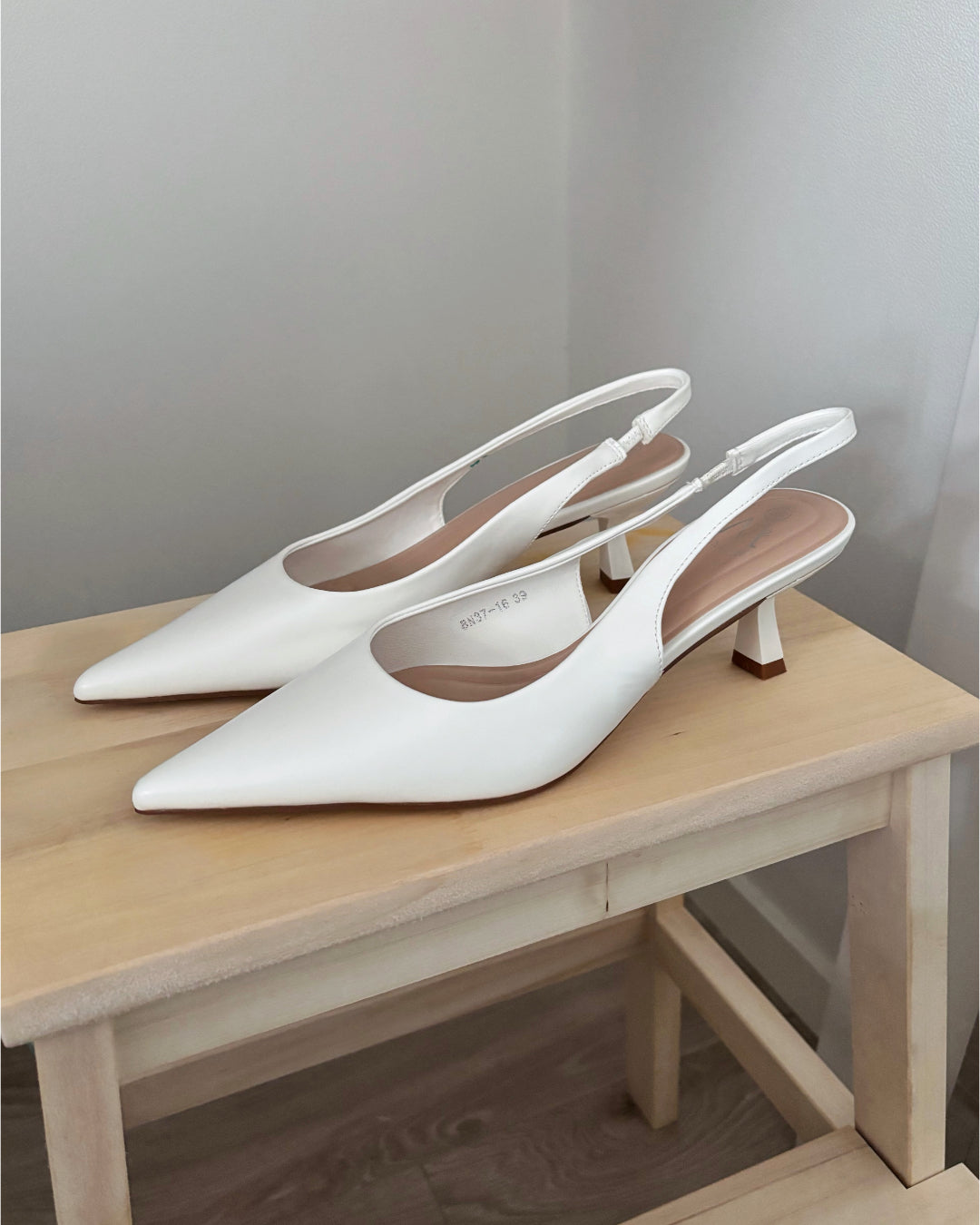 Hvide slingback-højhælede sko står på en lys træskammel i et hjørne af et rum. Skoene har en spids tå og lille hæl. Tekst: "8W37-1E 39".