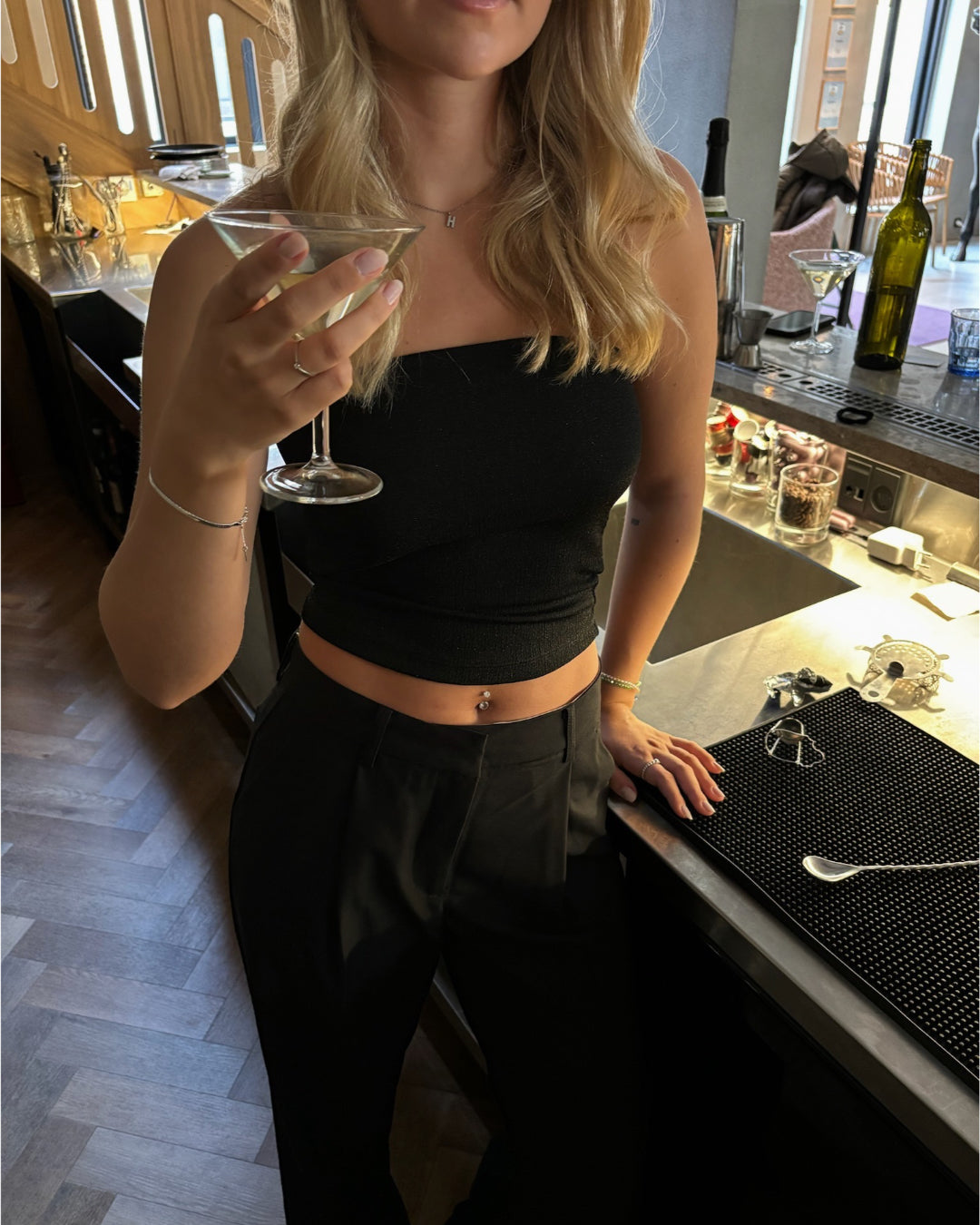 En kvinde i sort top holder et cocktailglas ved en bar. Detaljerede elementer som en flaske, glas og barudstyr er synlige i baggrunden.