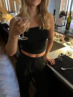 En kvinde i sort top holder et cocktailglas ved en bar. Detaljerede elementer som en flaske, glas og barudstyr er synlige i baggrunden.
