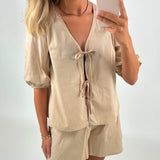 Kvinde holder en smartphone foran et spejl, iført en lys beige bluse med båndbinding og matchende shorts. Hun har lyst, bølget hår, guldøreringe og et enkelt armbånd.
