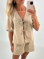 Kvinde holder en smartphone foran et spejl, iført en lys beige bluse med båndbinding og matchende shorts. Hun har lyst, bølget hår, guldøreringe og et enkelt armbånd.