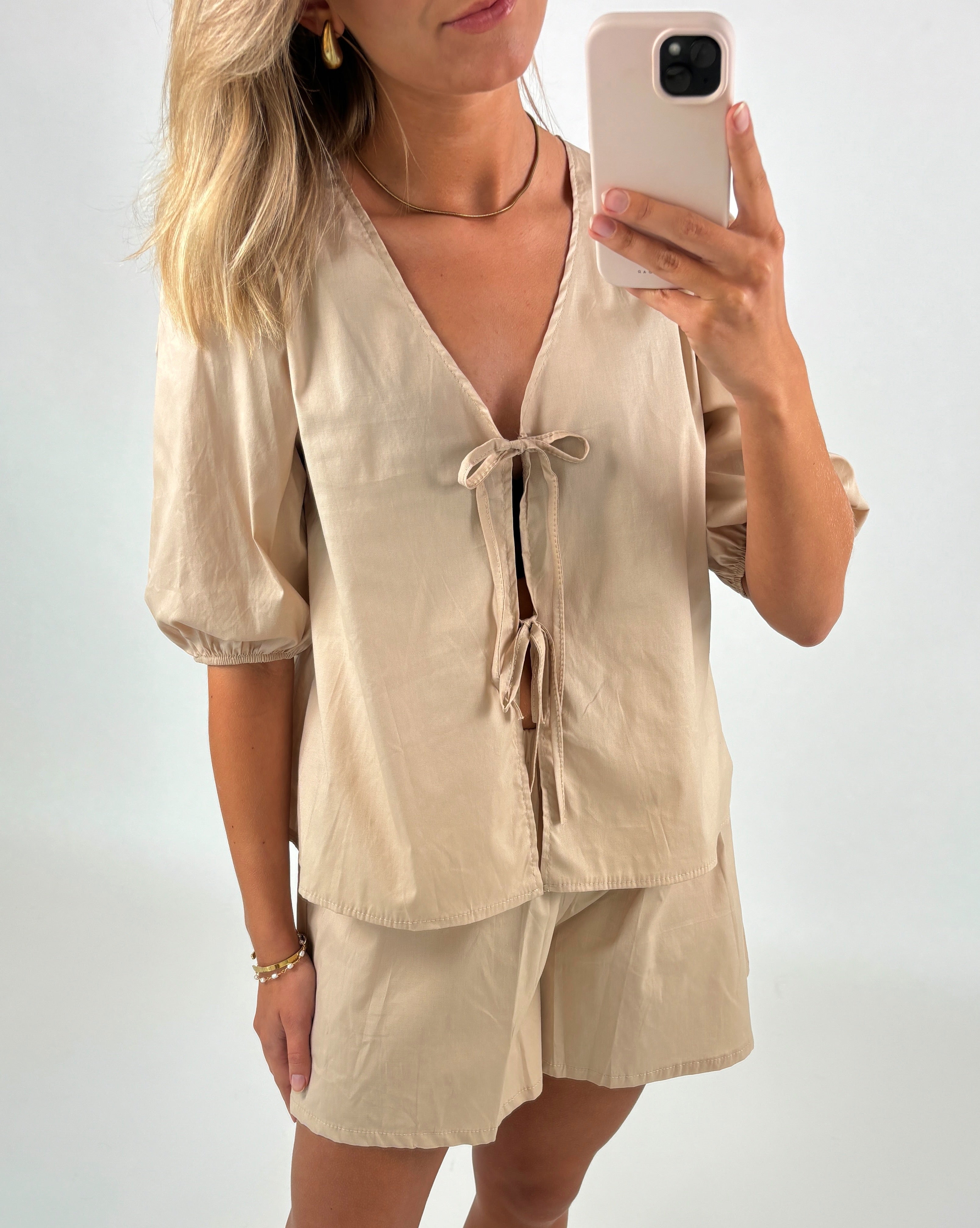 Kvinde holder en smartphone foran et spejl, iført en lys beige bluse med båndbinding og matchende shorts. Hun har lyst, bølget hår, guldøreringe og et enkelt armbånd.