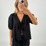 En person i sort bluse og shorts tager et spejlbillede med en smartphone. Personen står i et lyst rum og har skulderlangt blondt hår.