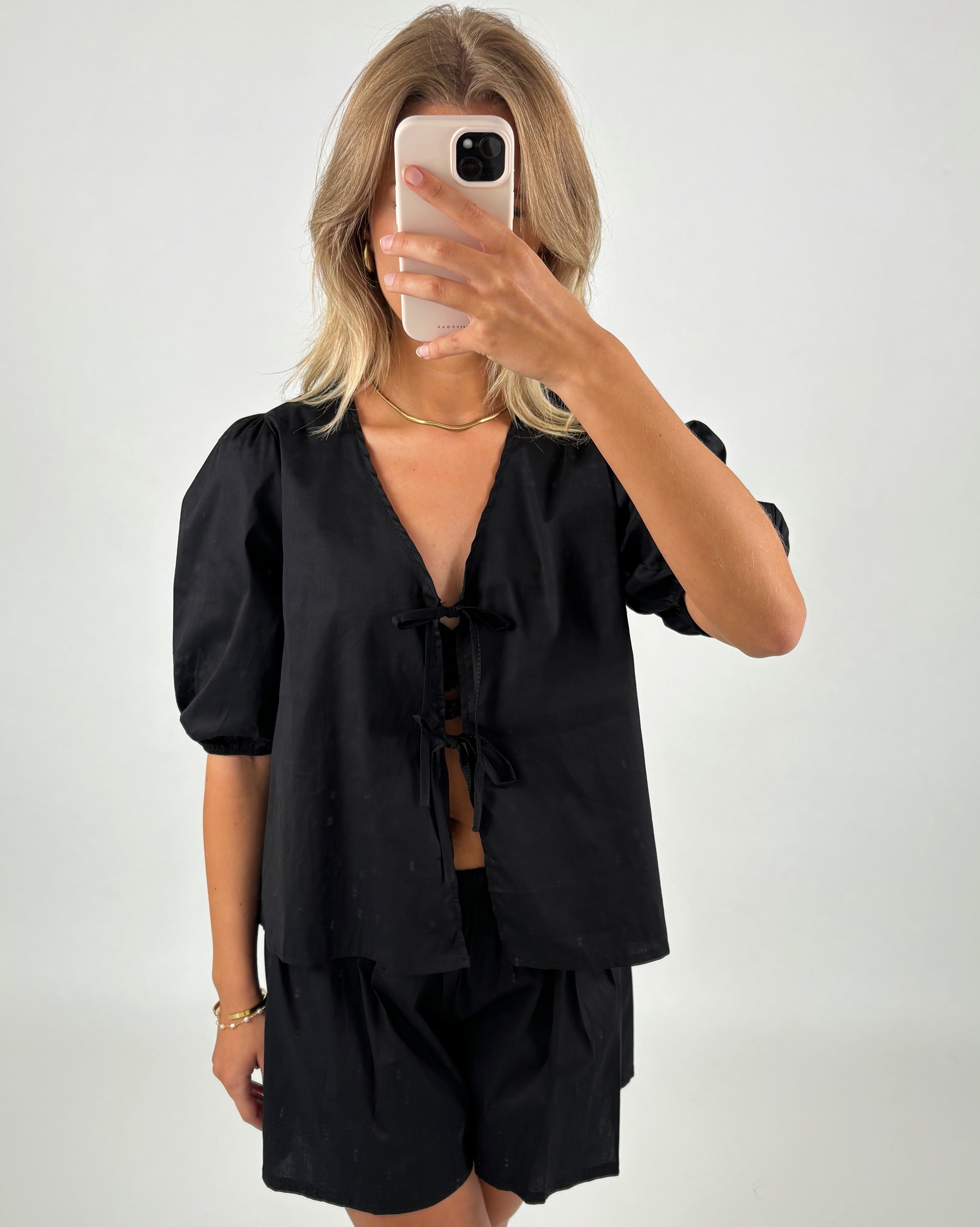 En person i sort bluse og shorts tager et spejlbillede med en smartphone. Personen står i et lyst rum og har skulderlangt blondt hår.