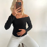 En person iført sort off-shoulder bluse og hvide bukser sidder på knæ og tager et spejlbillede med en telefon, i en lys, minimalistisk indretning.