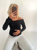 En person iført sort off-shoulder bluse og hvide bukser sidder på knæ og tager et spejlbillede med en telefon, i en lys, minimalistisk indretning.