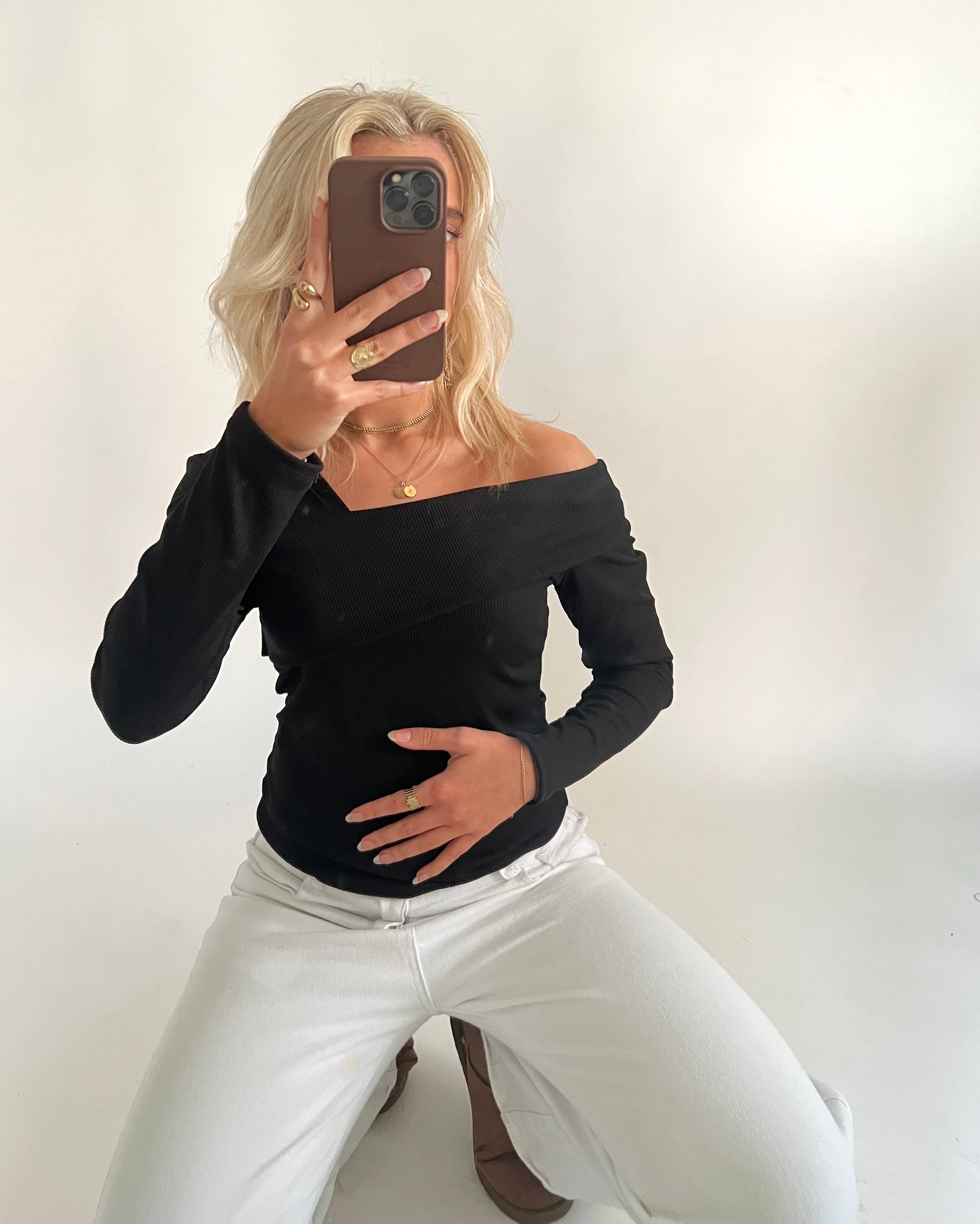 En person iført sort off-shoulder bluse og hvide bukser sidder på knæ og tager et spejlbillede med en telefon, i en lys, minimalistisk indretning.